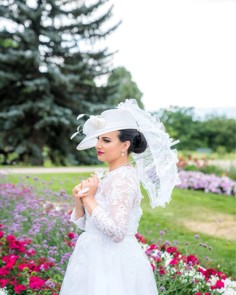 Bride in a wide-brim hat holding a bouquet in a flower garden, veil catching light